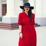 Young stylish woman wearing red maxi dress, black leather jacket and hat walking on the city street in autumn. Fall fashion, elegant look. Plus size model.